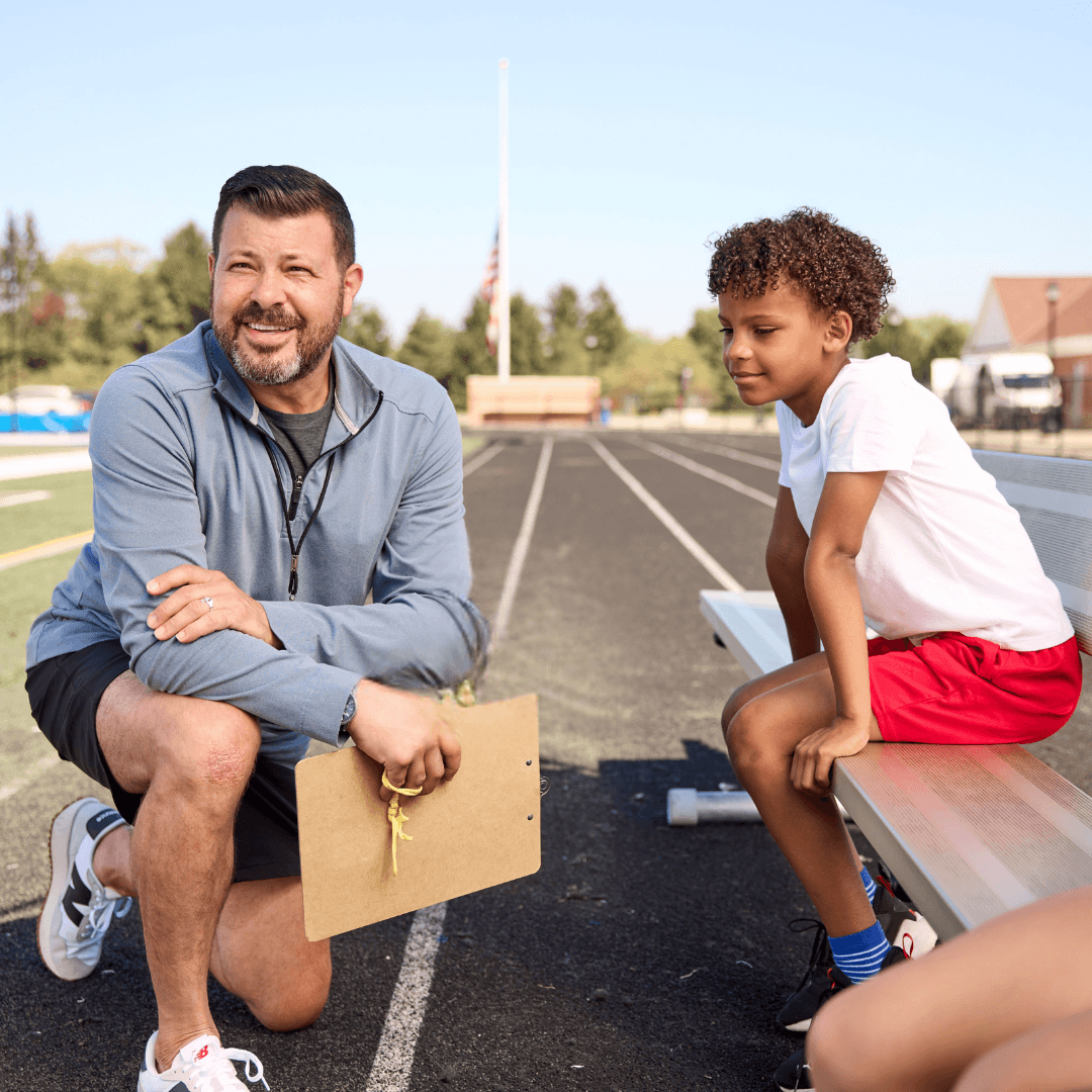 A coach kneels with a clipboard, talking to a young boy in sports attire on a track field bench.